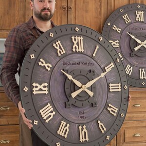 May include: A large round wall clock with a dark purple and black distressed finish. It features raised cream-colored Roman numerals, gold-toned hands, and the text "Enchanted Knights" and "Est. 1995" printed on the face. The clock has a wide border.