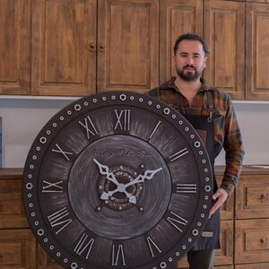 May include: Large, round, dark brown wall clock with Roman numerals and decorative hands. The clock face has the text "Quality Table Time" and "Sabne Ateliers". The clock is surrounded by small, evenly spaced circles.