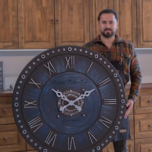 Könnte beinhalten: Große, runde, dunkelblaue und graue Wanduhr mit römischen Ziffern. Das Zifferblatt trägt den Text "Quality Takes Time" und "Kohne Atelier". Die Uhr wird von einer Person gehalten.