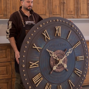 May include: Large, round wall clock with a dark brown frame and a blue-green center. Gold-colored Roman numerals and clock hands are visible. The clock has the words "Leap of Faith" on the face. The clock is being held by a person.