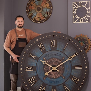 May include: Large, round wall clock with a dark brown frame, blue and brown face, and gold Roman numerals. The clock has the text "BOHEMART" and "BITLIS". Additional clocks are visible in the background.