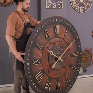 May include: Large, round wooden clock with Roman numerals and gold-colored hands. The clock face is brown with a dark brown border and the text "BOHEMART BITLIS". The clock is being held by a person.