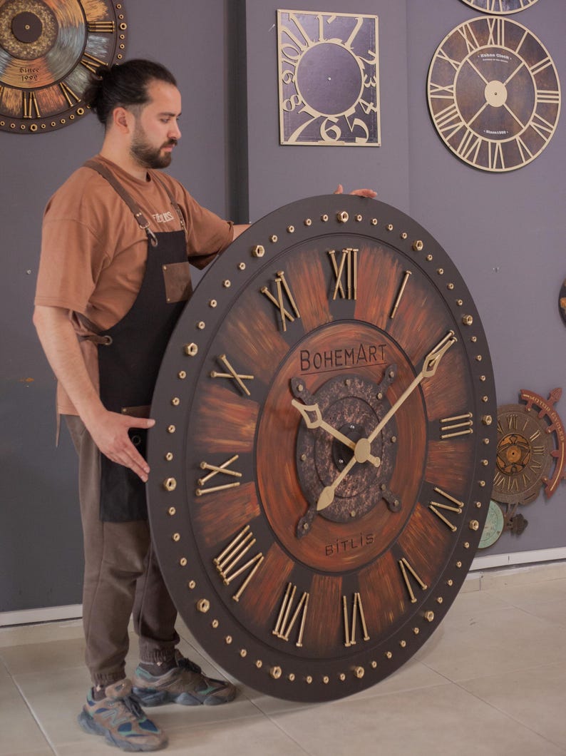 May include: Large, round wooden clock with Roman numerals and gold-colored hands. The clock face is brown with a dark brown border and the text "BOHEMART BITLIS". The clock is being held by a person.