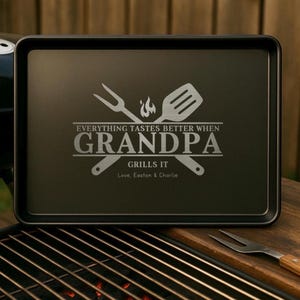 May include: A black rectangular grilling tray with the text "EVERYTHING TASTES BETTER WHEN GRANDPA GRILLS IT" and a fork and spatula design. The tray is on a wooden surface near a barbecue.