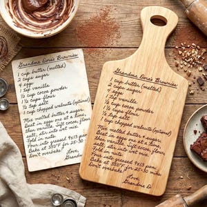 Puede incluir: Una tabla de cortar de madera y una tarjeta de receta con la receta manuscrita de "Brownies de la abuela Dora". La receta incluye ingredientes como mantequilla, azúcar, huevos y cacao en polvo. La tabla de cortar tiene un asa.