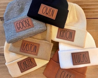 May include: A collection of six knit beanies in black, gray, beige, cream, and brown. Each beanie features a rectangular faux leather patch with a name engraved in black text: GOOSE, OWEN, KIPTON, EASTON, CARTER, LUCAS, and CLAY.