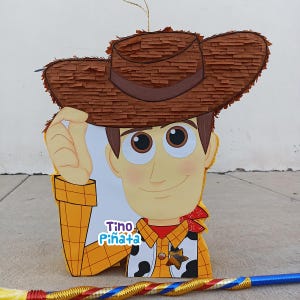 May include: A Woody-themed pi&ntilde;ata, featuring a brown cowboy hat, a yellow shirt with a sheriff's badge, and a red bandana. The pi&ntilde;ata has a smiling face and is made of paper. A colorful stick lies at the bottom.