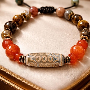 Puede incluir: Pulsera de cuentas con una cuenta cilíndrica central con un patrón geométrico en tonos azules, beige y blanco. La pulsera incluye cuentas naranjas, marrones y beige, con un cordón negro ajustable. Se muestra sobre una superficie crema.