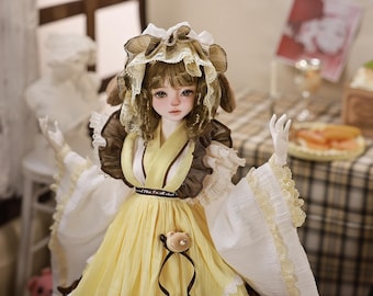May include: A detailed image of a doll with a yellow dress and a brown and white bonnet. The doll has a delicate face with rosy cheeks and is posed with its arms outstretched. The dress has a ribbon and a small bear decoration. The background is blurred.