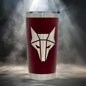 May include: A maroon tumbler featuring a silver geometric wolf head design. The cylindrical container has a metallic silver rim at the top and a matching silver base. A clear plastic lid is secured to the top of the vessel, which displays a sharp, angular, and stylized animal graphic.