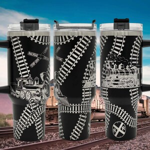 May include: Black travel mugs with a train theme. The mugs feature a black background with white train tracks and train illustrations. Each mug has a black handle and a silver lid. The mugs have the text "RAILROAD CROSSING" and "STOP LOOK LISTEN".