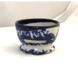 May include: A small, white ceramic egg cup with a blue and white floral pattern. The cup has a flared rim and a rounded base.