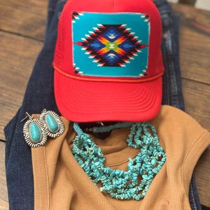 May include: A red trucker hat with a turquoise and geometric design patch, paired with a turquoise necklace, earrings, and a brown top. The hat is resting on a pair of blue jeans, creating a casual, southwestern-inspired ensemble.