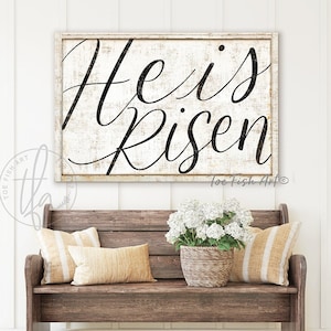 May include: A white wooden sign with black calligraphy that reads "He is Risen" with the artist's signature "toe fish Art" in the bottom right corner. The sign is hanging on a white wall above a wooden bench with white and tan striped cushions and a wicker basket filled with white flowers.