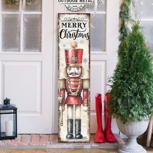 May include: A tall, rectangular vertical sign featuring a vintage-style illustration of a red and gold nutcracker soldier. The text "MERRY Christmas" is printed in black at the top. The sign has a weathered, distressed finish with snowflake accents.