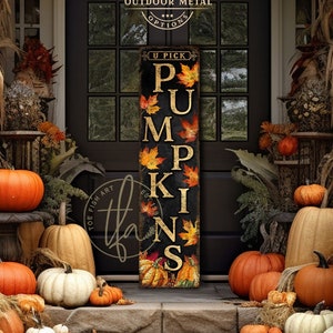 May include: A vertical rectangular sign with a black background featuring the text U PICK PUMPKINS in gold lettering, decorated with colourful autumn leaves and pumpkin illustrations. The sign is positioned in front of a dark door with glass panes.