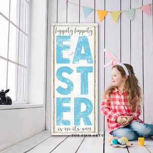 May include: A white wooden sign with blue text that reads "Hippity Hoppity Easter is on its way" with the words "Easter" in large, stylized letters. A young girl wearing bunny ears sits on a white wooden floor with a basket of Easter eggs.