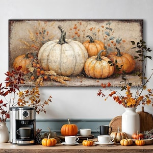 May include: A rectangular wall art print featuring a collection of pumpkins in shades of white and orange, surrounded by dried wheat and foliage. Below, a kitchen counter displays a coffee maker, white mugs, and several small orange pumpkins.