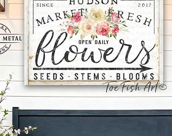 May include: A rectangular wall sign featuring the text "HUDSON MARKET FRESH flowers SINCE 2017 OPEN DAILY SEEDS STEMS BLOOMS" with a floral illustration of pink and yellow roses. The sign has a white background with black lettering and an arrow.