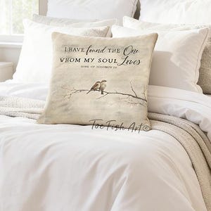 May include: A decorative pillow with the text "I have found the One whom my soul loves" and a drawing of two birds on a branch. The pillow is beige and sits on a white bed with white bedding. The room has two white bedside tables with lamps.