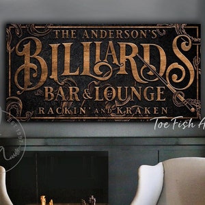 May include: A wooden sign with the text "The Anderson's Billiards Bar & Lounge Rackin' and Kraken" in a vintage style font. The sign is brown with gold lettering and has a dark background.