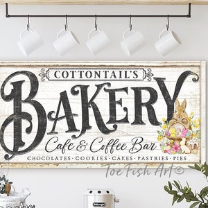 May include: A white sign with black lettering that reads "COTTONTAIL'S BAKERY Cafe & Coffee Bar CHOCOLATES COOKIES CAKES PASTRIES PIES Toe Fish Art" with a cartoon bunny holding a basket of Easter eggs.