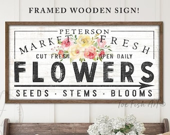 May include: A framed wooden sign with a white background and black text that reads "Peterson Market Fresh Cut Fresh Open Daily Flowers Seeds Stems Blooms". The sign is decorated with a watercolor illustration of pink, yellow, and white flowers.