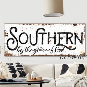 May include: White wooden sign with black lettering that reads "Southern by the grace of God" with a distressed look. The sign is hanging on a white wall above a white couch with black and white pillows.