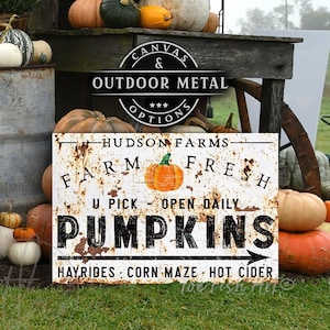 May include: A white metal sign with black text that reads "Hudson Farms Farm Fresh U Pick Open Daily Pumpkins Hayrides Corn Maze Hot Cider". The sign is propped up against a pile of pumpkins and gourds in front of a wooden table and a rusty metal barrel.
