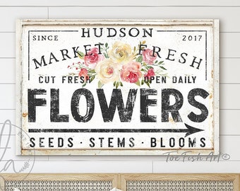 May include: A white sign with black text that reads "Hudson Market Fresh" with a floral design and the words "Cut Fresh" and "Open Daily" above the word "Flowers". The sign also says "Seeds, Stems, Blooms" at the bottom.