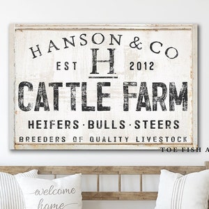 May include: A white sign with black text that reads "HANSON & CO EST H 2012 CATTLE FARM HEIFERS BULLS STEERS BREEDERS OF QUALITY LIVESTOCK TOE FISH ART". The sign is hanging on a white wall above a wooden bench.