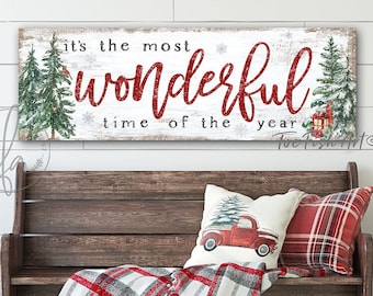 May include: A rectangular wall sign with a white wood-grain background featuring green pine trees, a red lantern, and the red glittery text "it's the most wonderful time of the year" displayed above a wooden bench with plaid pillows and a throw.