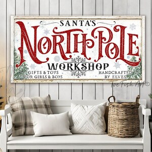 May include: A white sign with red lettering that reads "Santa's North Pole Workshop" with the text "Gifts & Toys for Girls & Boys" and "Handcrafted by Elves!"  The sign is hanging on a white wood wall with a white bench in front of it.