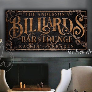 May include: A wooden sign with gold lettering that reads "The Anderson's Billiards Bar & Lounge Rackin' and Kraken" with a vintage style design. The sign is hanging on a dark wall above a pool table.