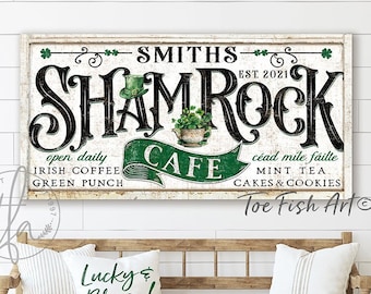 May include: A vintage-style sign for a cafe called "Smiths Shamrock Cafe" with green shamrock accents. The sign reads "open daily, Irish coffee, green punch, céad míle fáille, mint tea, cakes & cookies". The sign is hanging above a wooden bench with white pillows.