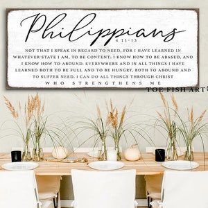 May include: White wooden sign with black text that reads "Philippians 4:11-13" and a Bible verse about contentment. The sign is hanging on a wall above a wooden table with white chairs.