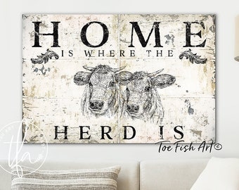 May include: A distressed white wood sign with black text that reads "Home is where the herd is" and a black and white illustration of two cows. The sign is a farmhouse style decor piece.