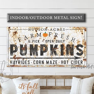 May include: Rustic metal sign with a white background and brown rust accents. The sign reads "Hudson Acres Farm Fresh U-Pick Open Daily Pumpkins Hayrides Corn Maze Hot Cider" with a small orange pumpkin graphic. The sign is hanging on a white wood wall above a wooden bench with two pillows.