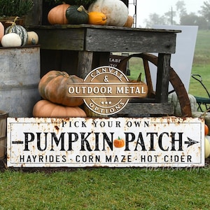 May include: A white sign with rusty edges reads "Pick Your Own - Pumpkin Patch - Hayrides Corn Maze Hot Cider". The sign is propped up against a wooden stand with pumpkins and gourds on top.