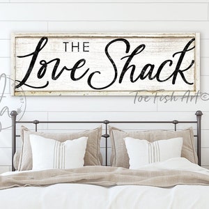 May include: A white wooden sign with black lettering that reads "The Love Shack" with the words "Toe Fish Art" in the bottom right corner. The sign is hanging above a bed with white bedding and tan pillows.