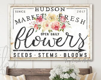 May include: White sign with black text that reads "Since 2017 Hudson Market Fresh Open Daily Flowers Seeds Stems Blooms". The sign has a distressed look and is decorated with pink and yellow flowers.