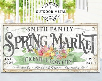 Könnte beinhalten: Ein rechteckiges Schild mit abgenutztem weißem Finish und dem Text Smith Family Spring Market in großen dunkelgrauen Buchstaben. Ein grünes Band zeigt Fresh Flowers mit bunten Blumenillustrationen und kleinerem Text.