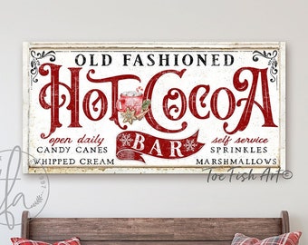 Puede incluir: Un letrero de estilo vintage con el texto "Old Fashioned Hot Cocoa Bar" en letras rojas sobre fondo blanco. El letrero también incluye el texto "Open Daily Candy Canes Whipped Cream" y "Self Service Sprinkles Marshmallows" en letras rojas más pequeñas. El letrero se muestra sobre un banco de madera con almohadas de cuadros rojos y blancos.