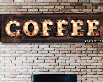 May include: A rustic brown metal sign with the word "COFFEE" in white letters with light bulbs around each letter. The sign is hanging on a brick wall above a grey sofa and a yellow armchair.