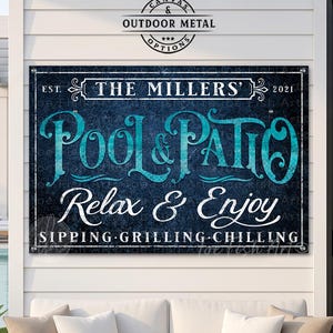 May include: A rectangular metal sign with a dark blue textured background and white and teal text that reads: EST. THE MILLERS' 2021, POOL & PATIO, Relax & Enjoy, SIPPING GRILLING CHILLING. The sign is mounted on a white wall above a white sofa.