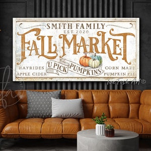 May include: A white sign with the text "Smith Family Fall Market Est. 2020" in brown lettering. The sign also includes the text "Hayrides Apple Cider U Pick Pumpkins Corn Maze Pumpkin Pie" in black lettering.