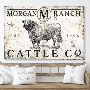 May include: A rustic white sign with black text and a black and white illustration of a bull. The sign reads "Morgan M Ranch Est. 1998 Cattle Co. Toe Fish Art".