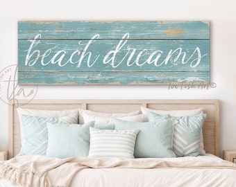 Może przedstawiać: Jasnoniebieski drewniany znak z napisem "beach dreams" w białym kolorze. Znak jest postarzony i ma rustykalny wygląd. Znak wisi nad łóżkiem z jasnoniebieskimi i białymi poduszkami.