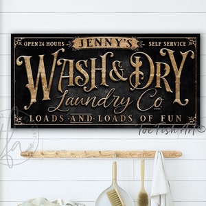 May include: A black and gold sign with the text "Jenny's Wash & Dry Laundry Co. Loads and Loads of Fun" on a white wood background.