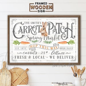 May include: A framed wooden sign with a white background and black lettering that reads "The Smith's Carrot Patch Spring Market". The sign also includes a cartoon bunny rabbit, carrots, and the text "Hop This Way", "Open Daily", "Carrots .25¢", "Lettuce", and "Fresh & Local - We Deliver".